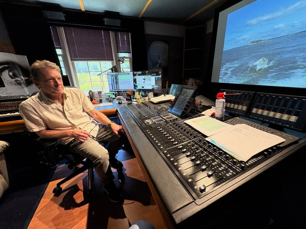 Tom Myers sits in his recording studio, looking at a large mixing console and a screen displaying an ocean scene with a boat.