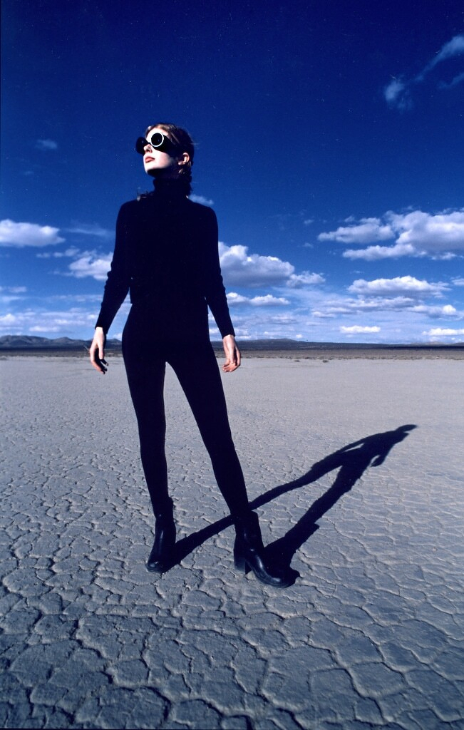Miranda Lee Richards  dressed in all black with large sunglasses stands on a cracked dry desert floor under a bright blue sky with scattered clouds.